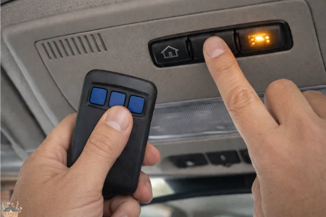 Hand holding a garage remote close to vehicle overhead console while pressing built-in control button with indicator light illuminated